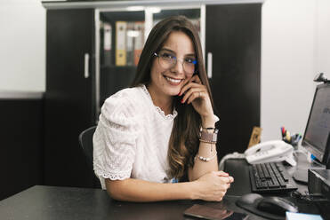 Geschäftsfrau lächelt, während sie mit dem Kopf in der Hand im Büro sitzt - DSIF00217