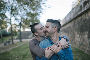 Glücklicher Mann, der seinen Freund auf die Wange küsst, während er sich von hinten im öffentlichen Park umarmt - GRCF00517