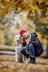 Smiling teenage girl with arm around on poodle crouching on road at park - JATF01273