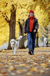 Smiling teenager girl walking with dog on road at park - JATF01278