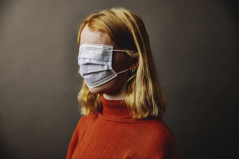 Blond girl wearing protective face mask on face while standing against gray background - JATF01287