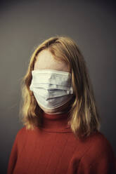 Teenage girl covering entire face with protective face mask while standing against gray background - JATF01288