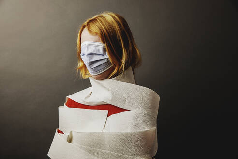 Teenager-Mädchen in Toilettenpapier eingewickelt und Gesicht mit Schutzmaske vor grauem Hintergrund stehend - JATF01290