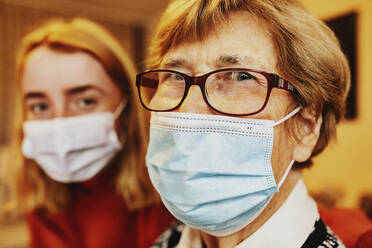 Grandmother wearing face mask and eyeglasses staring while sitting by granddaughter at home - JATF01296