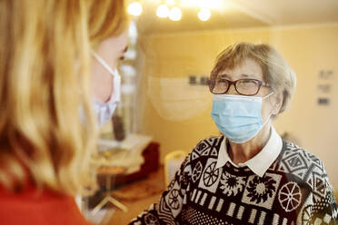 Grandmother wearing face mask looking at granddaughter while standing by glass at home during Covid-19 - JATF01298