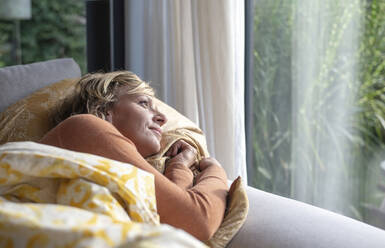 Reife Frau schaut durch das Fenster, während sie zu Hause auf dem Sofa liegt - BFRF02308
