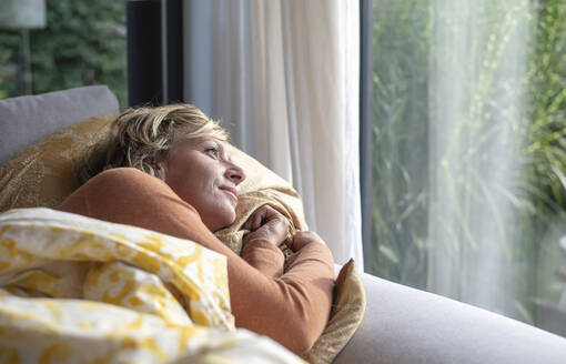 Mature woman looking through window while lying on sofa at home - BFRF02308
