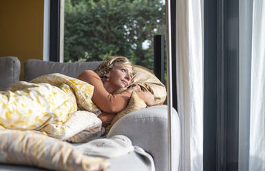 Nachdenkliche Frau schaut durch das Fenster, während sie zu Hause auf dem Sofa liegt - BFRF02309
