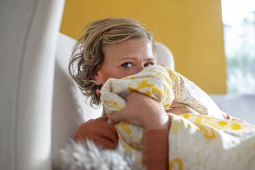 Frau bedeckt ihr Gesicht mit einer Decke, während sie zu Hause auf dem Sofa sitzt - BFRF02312