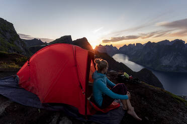 Frau bewundert die Aussicht, während sie im Lager auf dem Berg Reinebringen sitzt, Lofoten, Norwegen - MALF00190