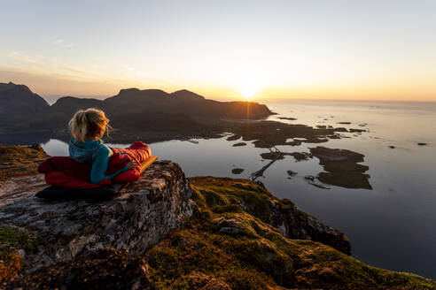 Wanderer bewundert die Aussicht auf den Sonnenuntergang, während er im Schlafsack auf dem Volandstinden, Lofoten, liegt - MALF00248