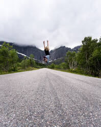 Frau mit erhobener Hand springt auf der Straße gegen einen Berg auf den Lofoten, Norwegen - MALF00258