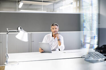 Nachdenklicher Geschäftsmann mit digitalem Tablet, der wegschaut, während er im Büro sitzt - PESF02251
