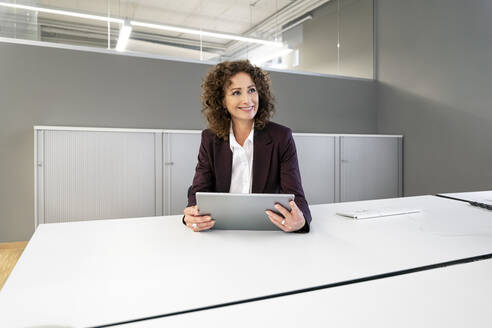 Geschäftsfrau mit digitalem Tablet, die wegschaut, während sie im Büro sitzt - PESF02257