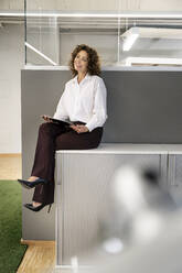 Nachdenkliche Geschäftsfrau mit digitalem Tablet, die wegschaut, während sie auf dem Tisch im Büro sitzt - PESF02261