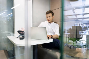 Junger Geschäftsmann arbeitet am Laptop, während er am Schreibtisch im Büro sitzt - PESF02276