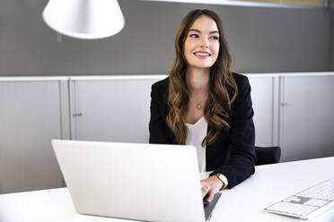 Smiling businesswoman with laptop looking away while sitting at office - PESF02288