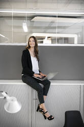 Geschäftsfrau mit Laptop, die wegschaut, während sie auf einem Tisch im Büro sitzt - PESF02289