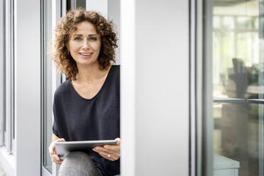 Businesswoman smiling while using digital tablet sitting at office door - PESF02301