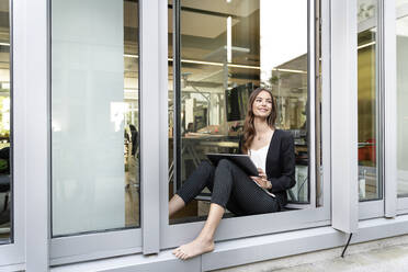 Lächelnde Geschäftsfrau mit digitalem Tablet, die wegschaut, während sie sich an die Bürotür lehnt - PESF02305