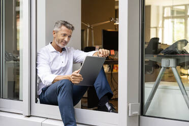 Lächelnder Geschäftsmann, der ein digitales Tablet benutzt, während er an der Bürotür sitzt - PESF02307