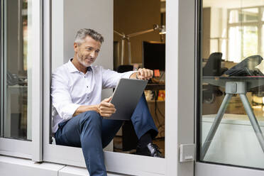 Smiling businessman using digital tablet while sitting at office door - PESF02307