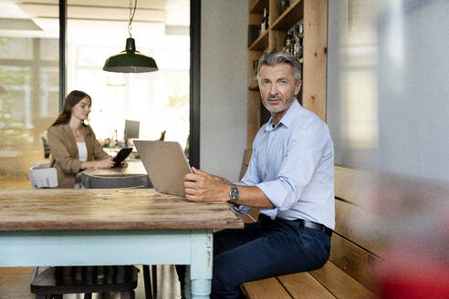 Geschäftsmann, der einen Laptop benutzt, während er mit einem Kollegen im Hintergrund im Büro sitzt - PESF02316