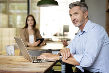 Mature businessman looking away while sitting with colleagues in background at office - PESF02320