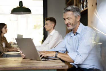 Älterer Geschäftsmann, der am Laptop arbeitet, während er mit Kollegen im Hintergrund im Büro sitzt - PESF02322