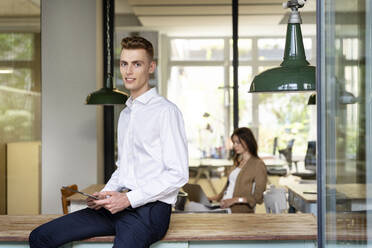 Junger Unternehmer, der ein digitales Tablet benutzt, während er auf einem Tisch mit einem Kollegen im Hintergrund im Büro sitzt - PESF02328