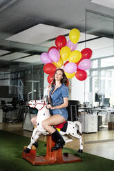 Young businesswoman holding multi colored balloon sitting on rocking horse at office - PESF02330