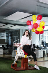 Junge Geschäftsfrau mit buntem Ballon sitzt auf Schaukelpferd im Büro - PESF02334