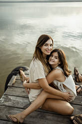 Mother and daughter embracing each other while sitting at jetty against lake - RCPF00319