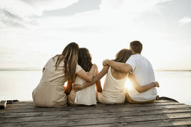 Parents and children with arms around sitting on jetty looking at view against sky - RCPF00337