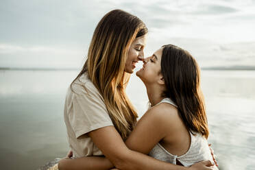 Mother and daughter nuzzling nose by lake against sky - RCPF00343