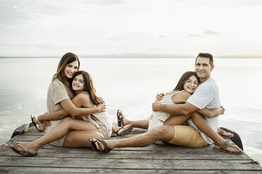 Töchter, die ihre Eltern umarmen, während sie auf dem Steg am See sitzen, gegen den Himmel - RCPF00344