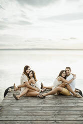 Töchter, die Mutter und Vater umarmen, während sie auf dem Steg am See sitzen, gegen den Himmel - RCPF00346