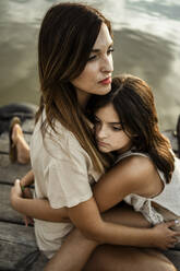 Daughter embracing thoughtful mother while sitting on jetty - RCPF00347