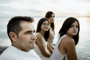 Father sitting with daughters and mother at jetty against lake - RCPF00357