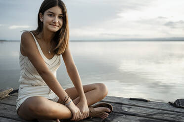 Smiling girl with cross legged sitting on jetty against sky and lake - RCPF00367