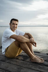 Smiling mature man sitting on jetty against lake - RCPF00369