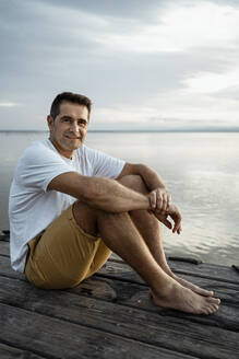 Lächelnder reifer Mann sitzt auf einem Steg am See - RCPF00369