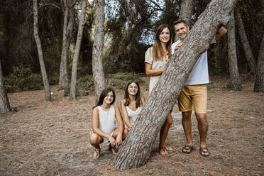 Eltern mit Töchtern stehen hinter einem Baumstamm im Wald - RCPF00379