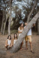 Mutter und Vater mit Töchtern hinter einem Baumstamm im Wald - RCPF00380