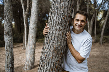 Smiling man embracing tree trunk in forest during vacation - RCPF00387