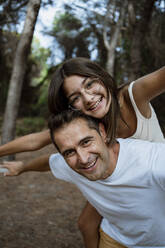 Father giving piggyback ride to daughter with arms outstretched in forest during vacation - RCPF00396