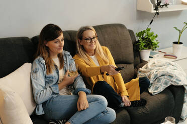 Smiling mother and daughter watching TV while sitting on sofa at home - ERRF04705