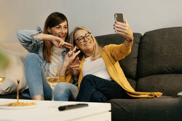 Lächelnde Mutter und Tochter, die ein Friedenszeichen machen, während sie ein Selfie mit dem Smartphone zu Hause machen - ERRF04709