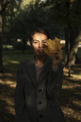 Female entrepreneur holding dry leaf while standing in park during autumn - VPIF03227