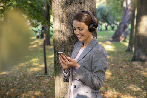 Lächelnde Geschäftsfrau hört Musik über Kopfhörer, während sie ein Smartphone im Park benutzt - VPIF03231